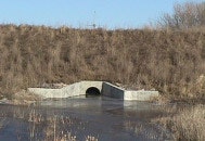 A concrete culvert with water flowing through it, surrounded by grasses and vegetation under a clear blue sky.
