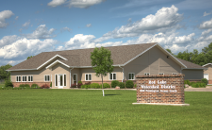 A single-story brick building with a gabled roof, surrounded by grass, and a sign in front on a sunny day.