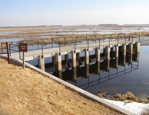 Concrete levee with metal railings over calm water, sign reads "Farmer's Pool," surrounded by grassy area.