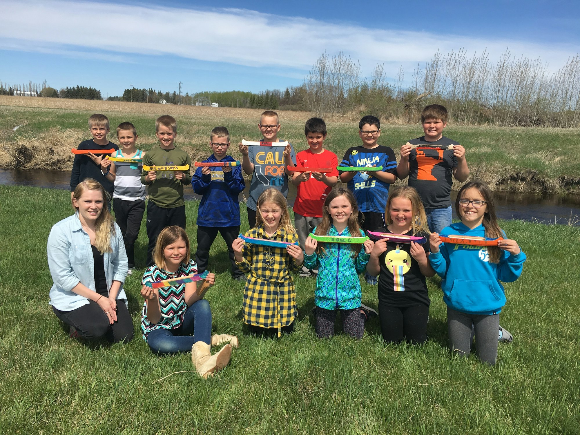A group of children and one adult outside, holding colorful wooden planes on a grassy field by a stream.