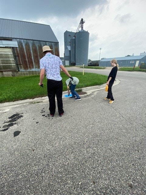 Three people on a rural road, one painting on the ground with a blue roller near industrial buildings.