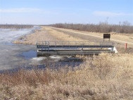 A small water control structure with a surrounding grassy field and a distant tree line under a clear sky.