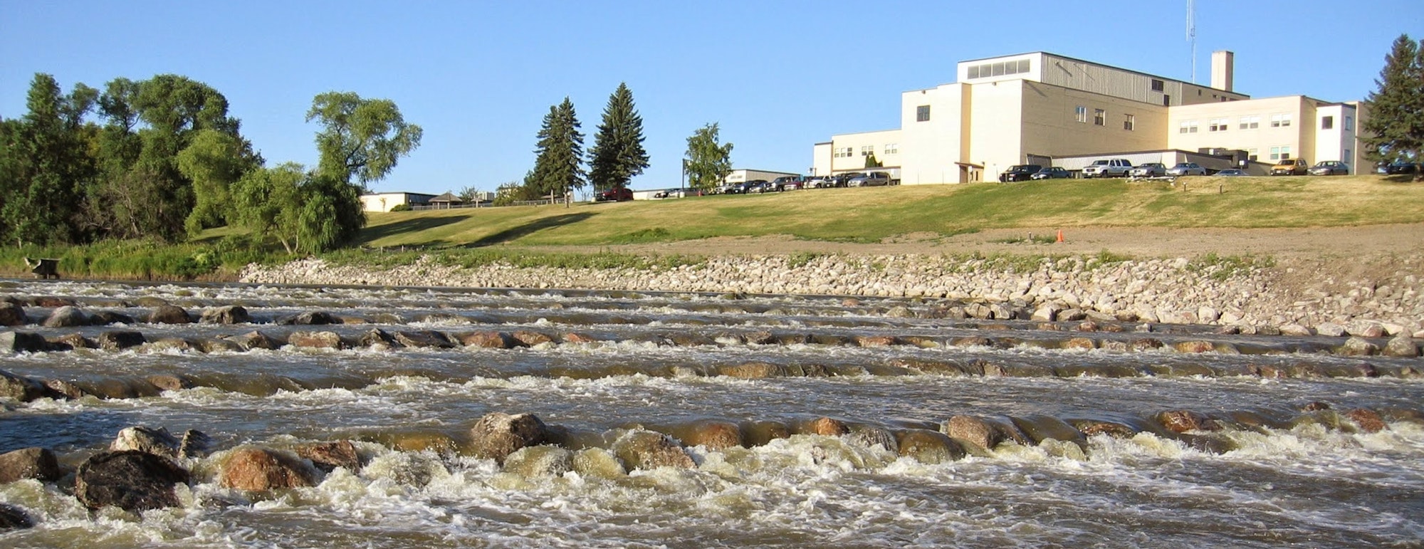 A building complex near a river with rocks, surrounded by grassy areas and trees under a clear blue sky.