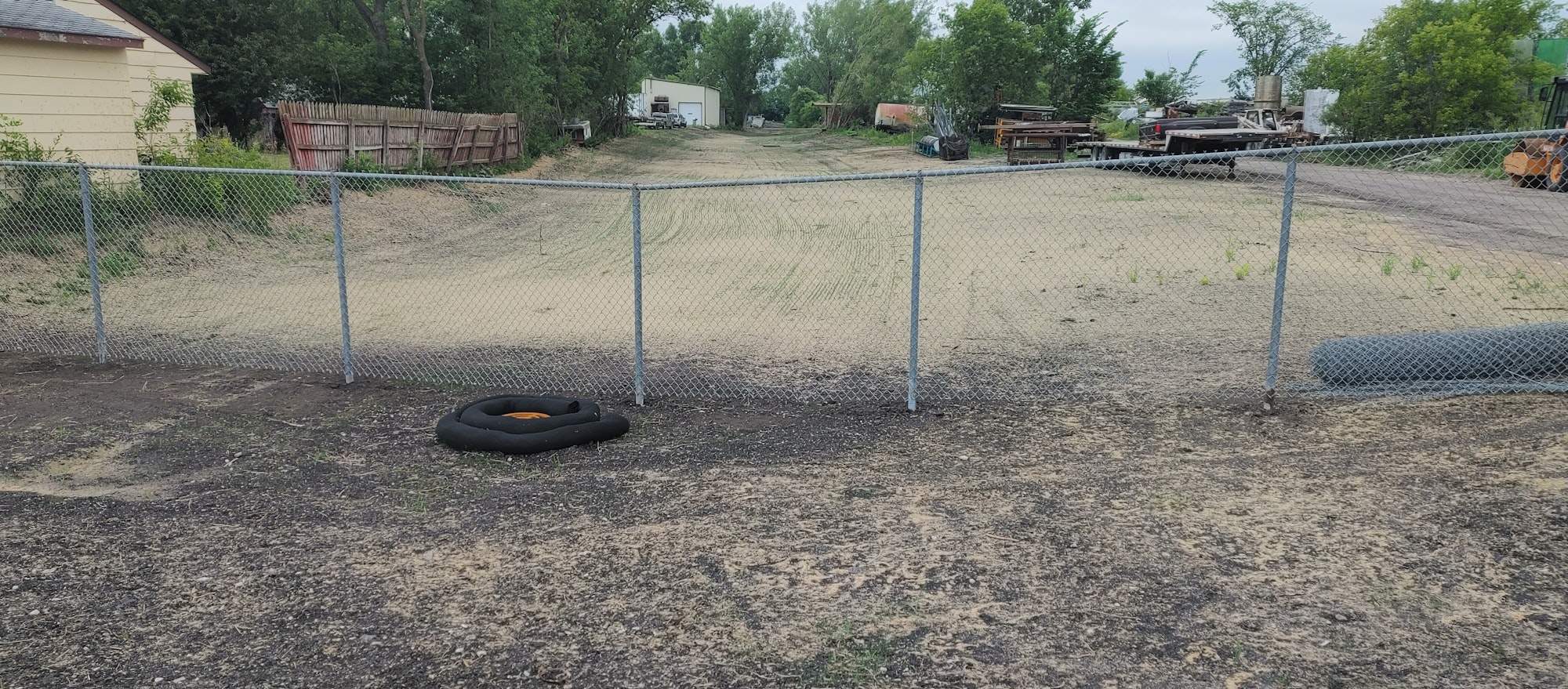 A chain-link fence with a coiled hose in front, leading to a yard with machinery and trees.