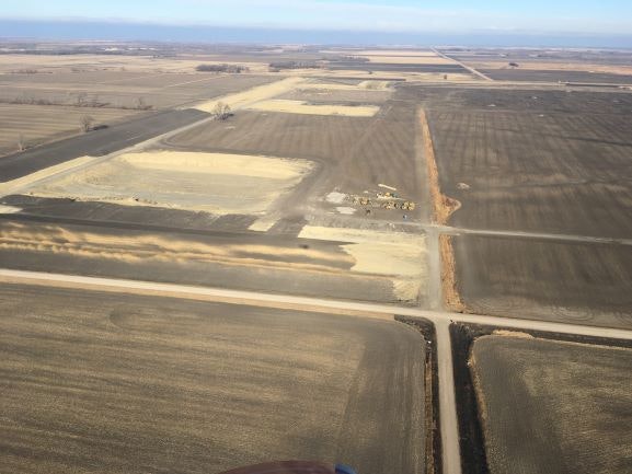 Aerial view of construction work on a rural landscape with roads, fields, and earth-moving equipment.