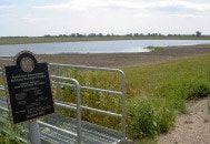 A small gate and plaque near a grassy area with a pond, under a partly cloudy sky.