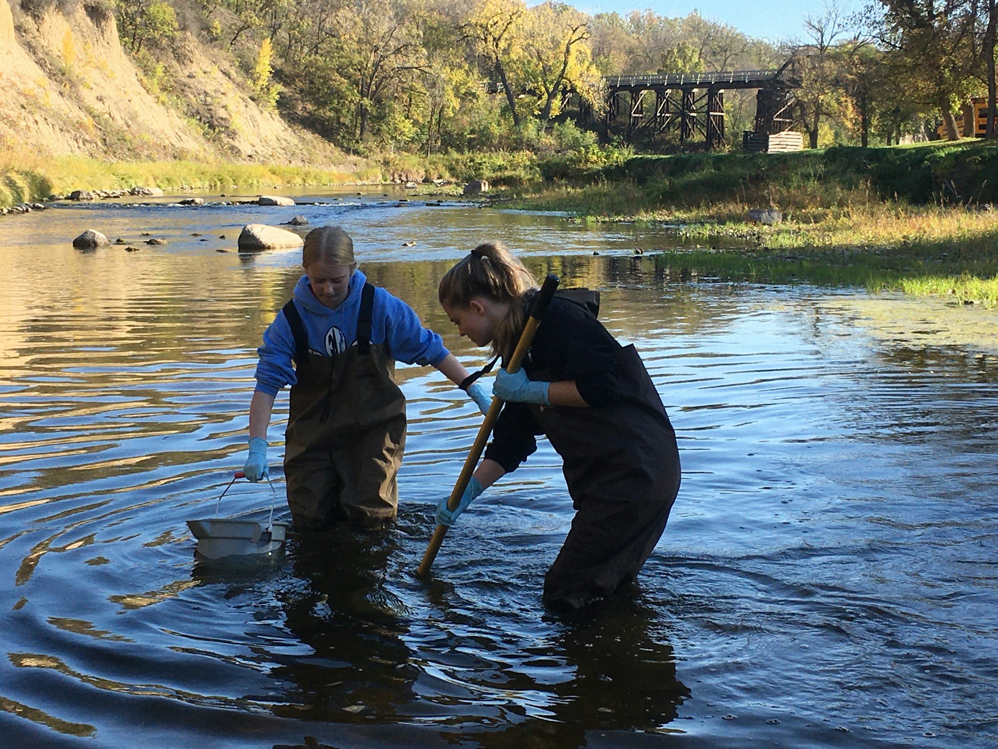 Two people in waders collect water samples in a river surrounded by trees and a bridge.