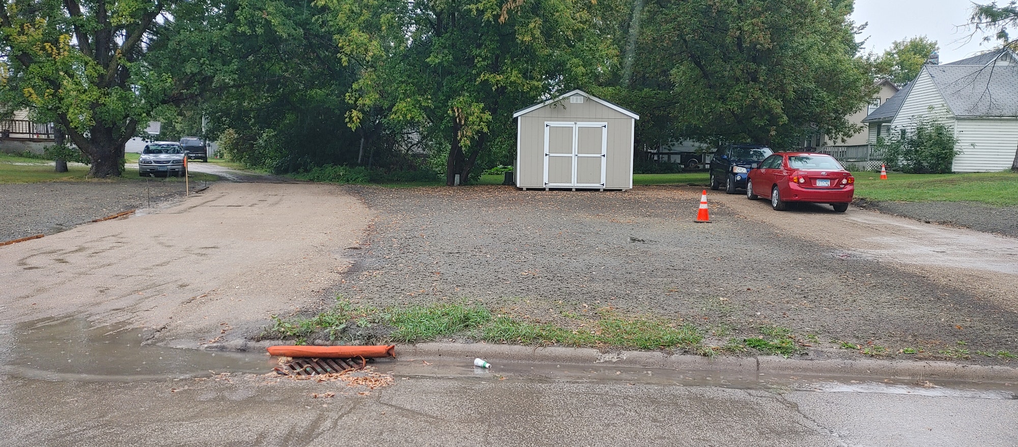 Paved area with a shed, cars, trees, and traffic cones. Curbside drain with water and a bottle.