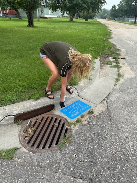 Person placing a blue sign saying "NO DUMPING - DRAINS TO WATERWAYS" on the curb near a storm drain on a street.