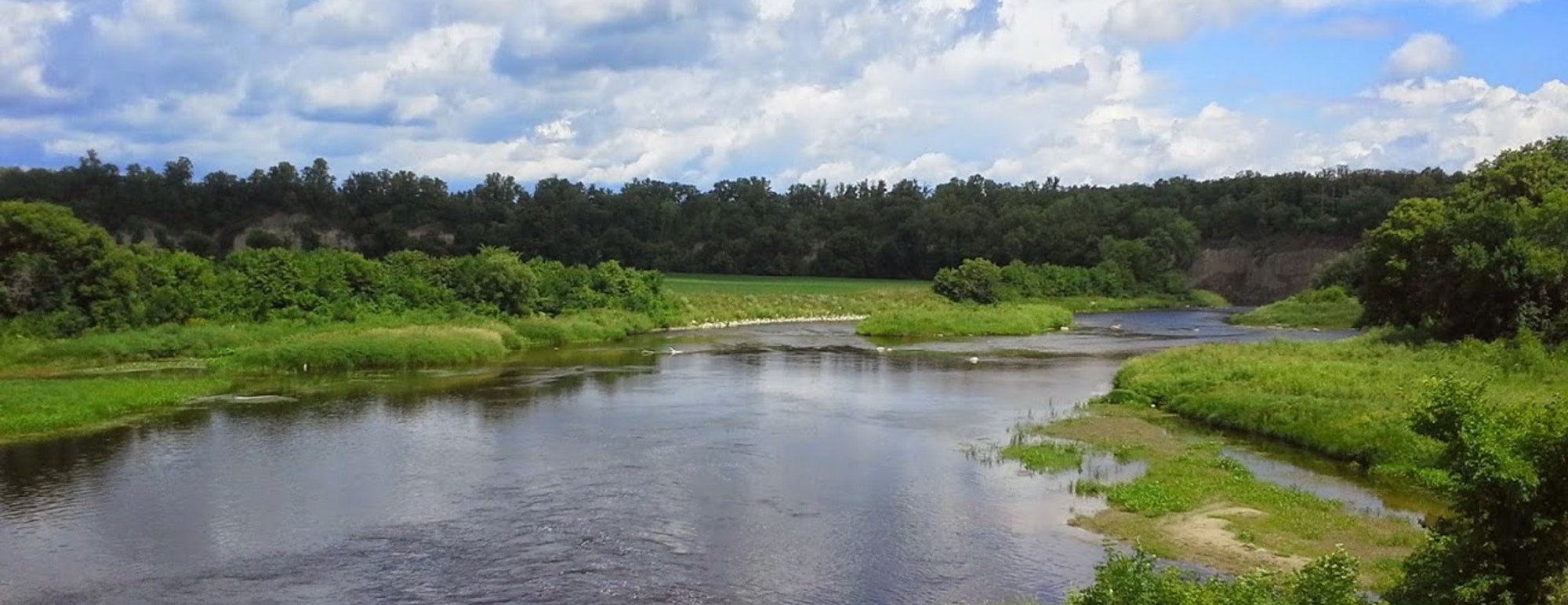 A serene river flows through lush greenery and trees under a partly cloudy blue sky.