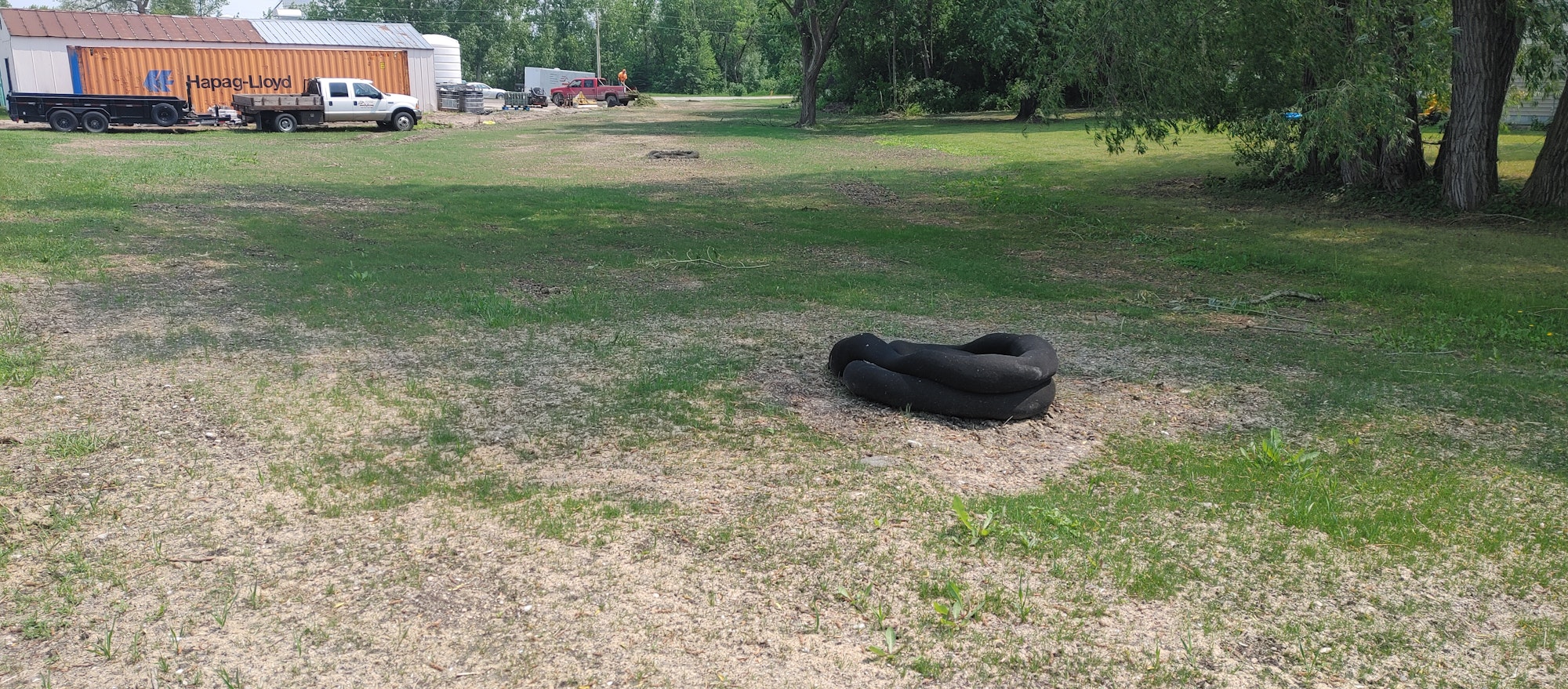 A grassy area with a black coiled object in the foreground and vehicles and containers in the background.