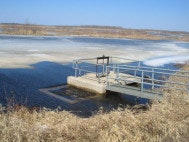 A small water control structure with a metal platform is surrounded by icy water and dry grass in a rural area.