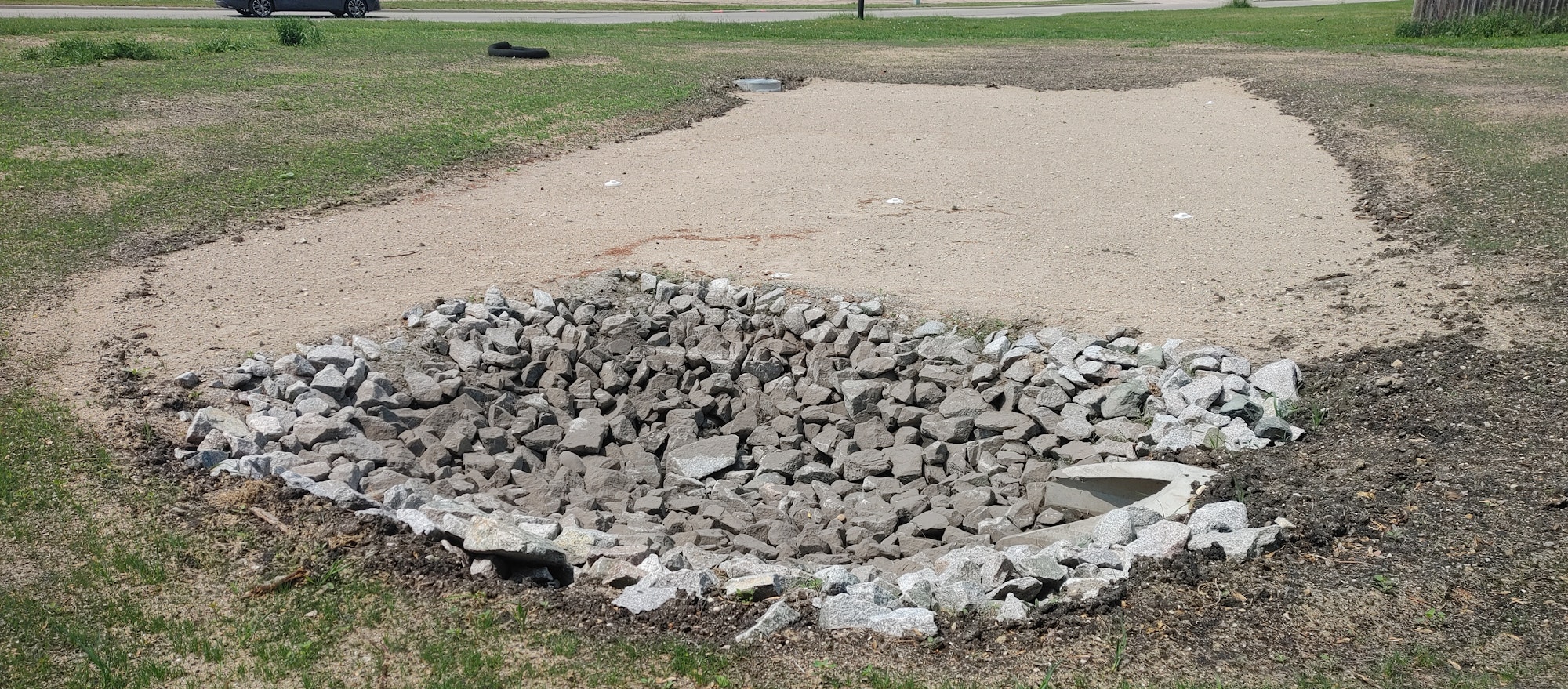 Rock drainage pit on a sandy area, surrounded by grass, with a roadway in the background.