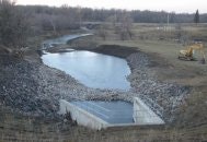 A river flows into a constructed inlet with concrete walls, surrounded by stones, in a rural landscape with bare trees and earthmoving equipment.