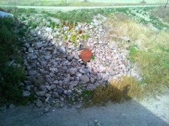 A pile of rocks with scattered grass and a small red bucket in the center, partially in shadow.