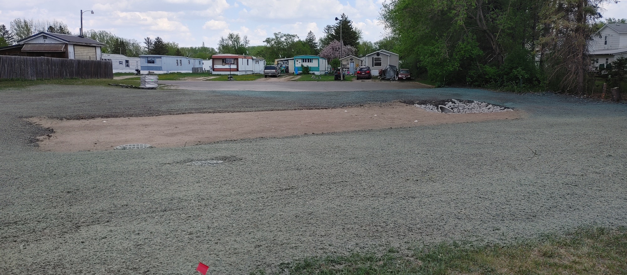 Open area with dirt, some grass, mobile homes in the background, and trees surrounding the scene.