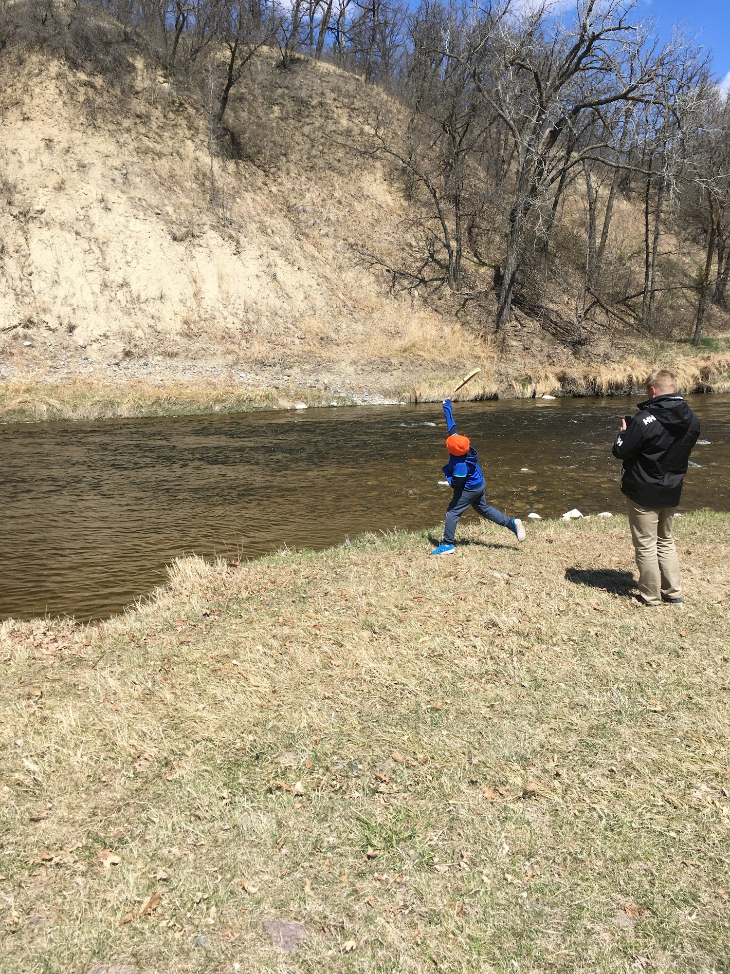 Two people by a riverbank; one in blue jumps mid-air, the other stands nearby. Sparse trees and grass surround them.