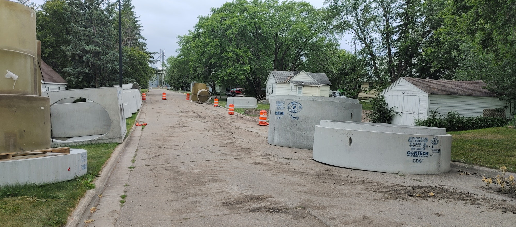 Street with construction, orange cones, large concrete pipes, and trees.