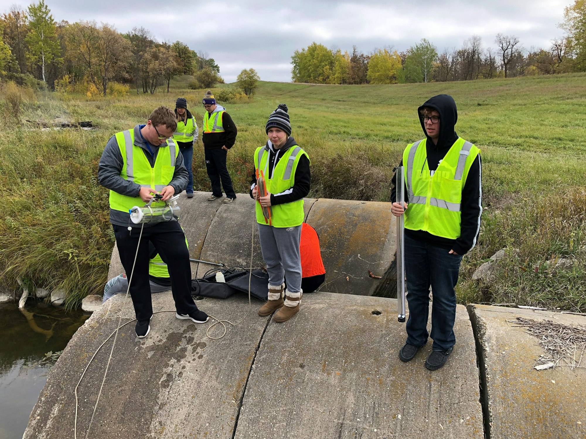 People wearing reflective vests conducting a field activity on a concrete structure near a small water body.