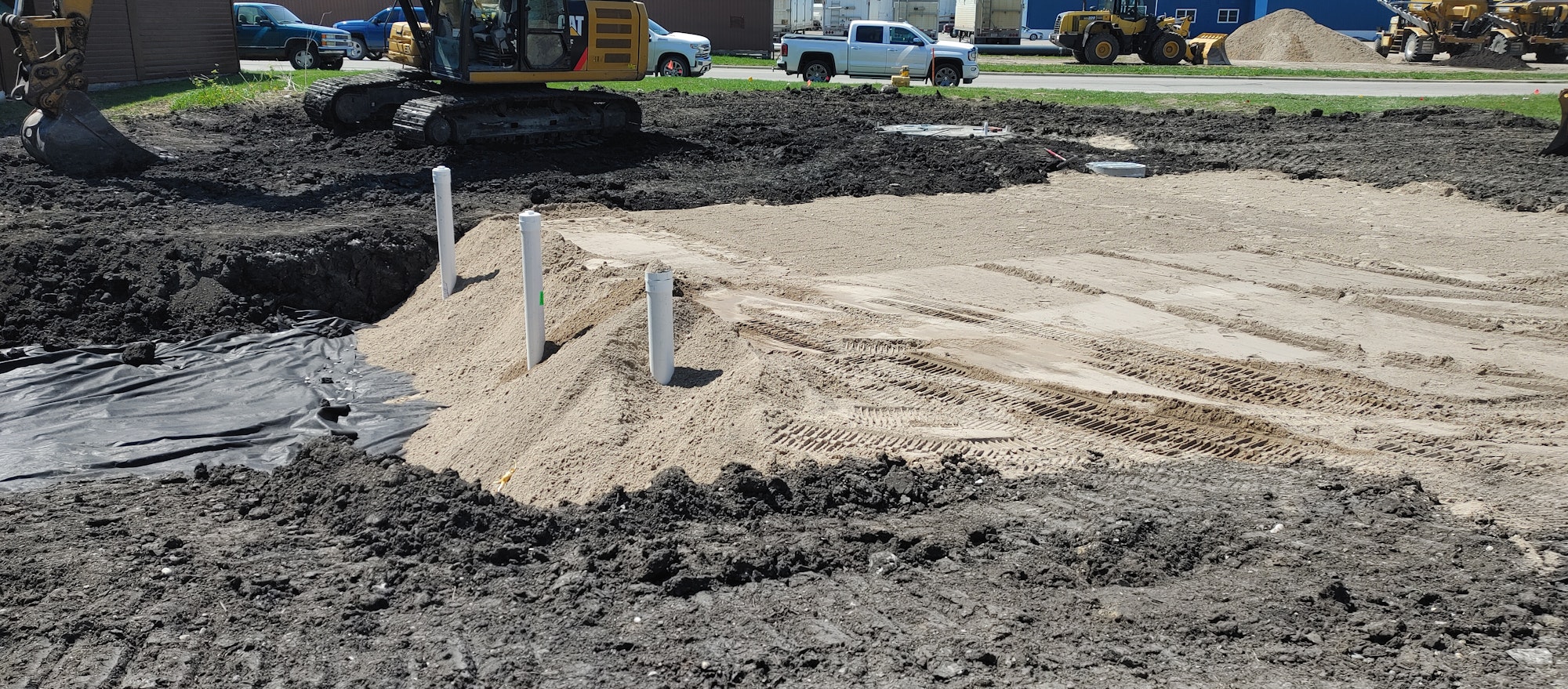 Construction site with an excavator, soil, sand, pipes, and tire tracks. Vehicles and buildings in the background.