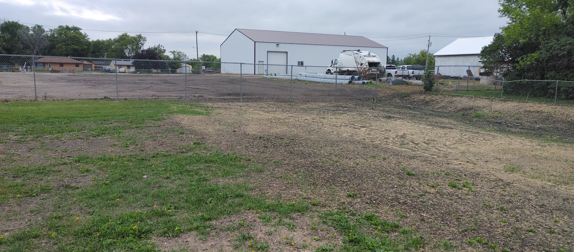 Fenced area with grassy foreground, industrial building, truck, and houses in the background. Overcast sky.