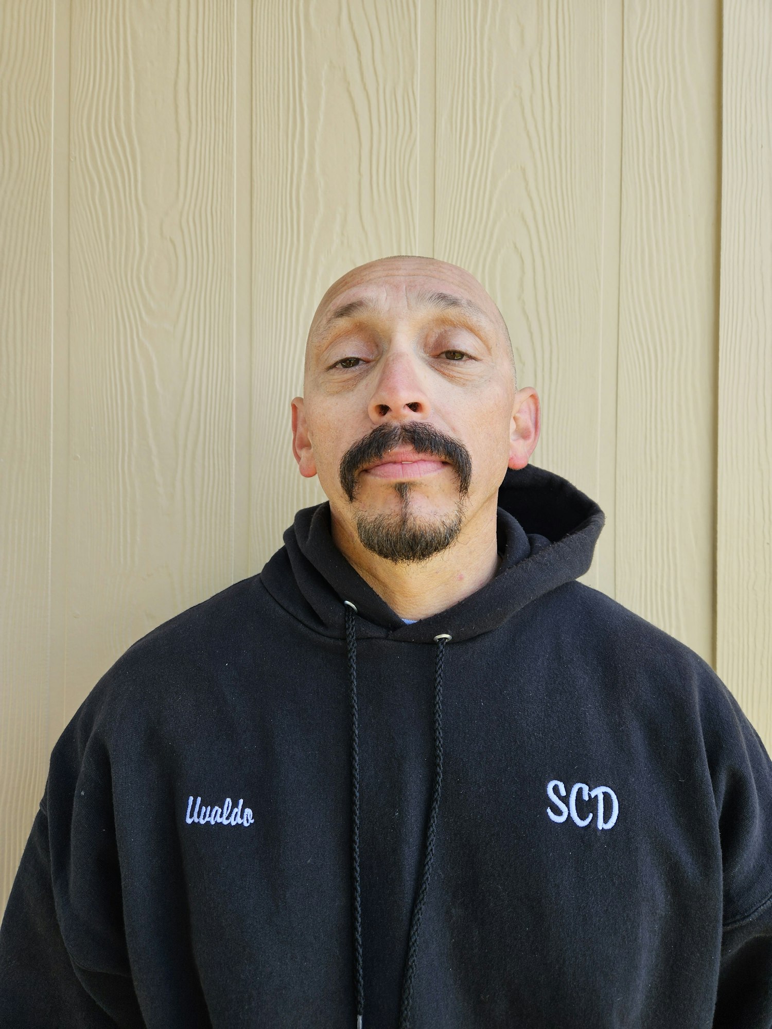 A person wearing a black hoodie with white lettering, standing against a light-colored wooden wall.