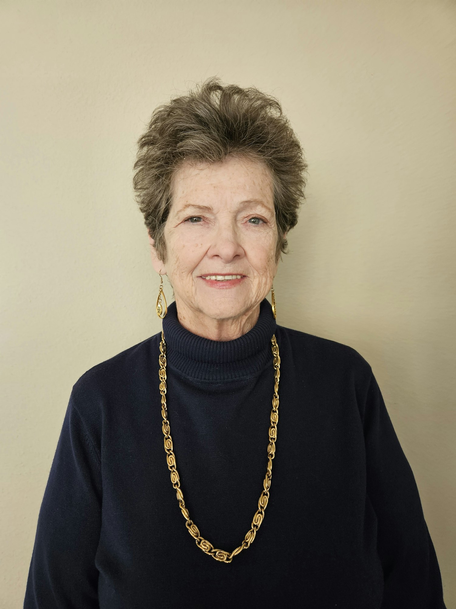 A smiling elderly woman wearing a navy sweater and gold necklace, photo rotated 90 degrees counterclockwise.