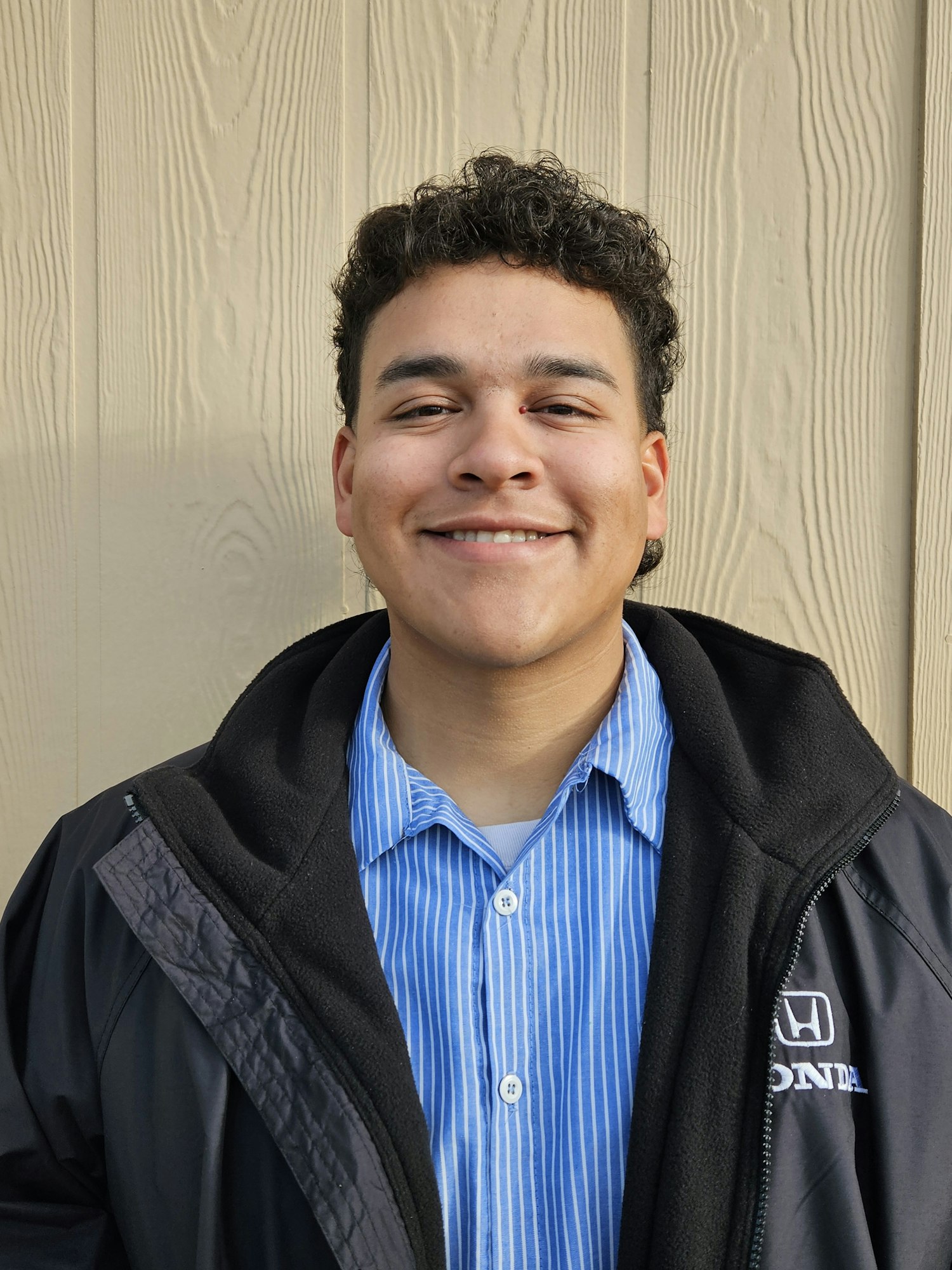 A person smiling, wearing a blue striped shirt under a black jacket, against a wooden background.