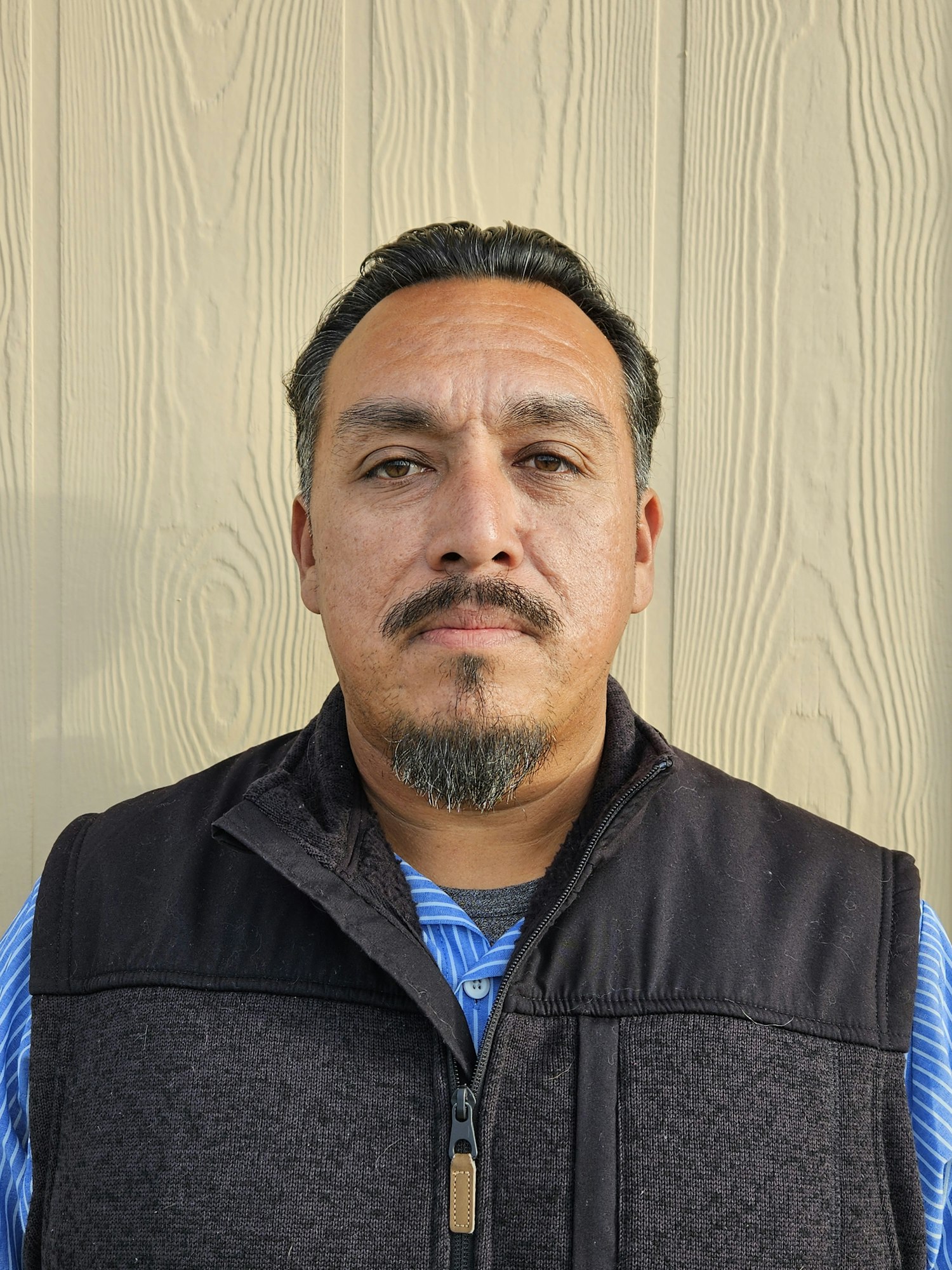 A man with a goatee and mustache is wearing a black vest over a striped shirt, standing against a beige wooden background.