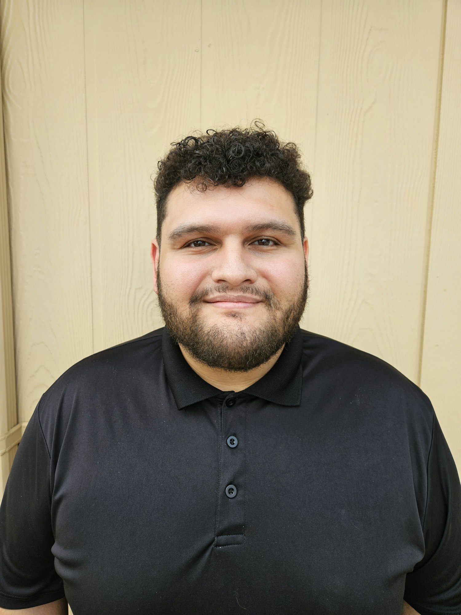 A person with curly hair and a beard, wearing a black polo shirt, standing against a light-colored wall.