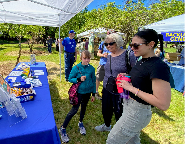 People at an outdoor event with informational booths, flyers, and promo items on a table, under a tent on a sunny day.