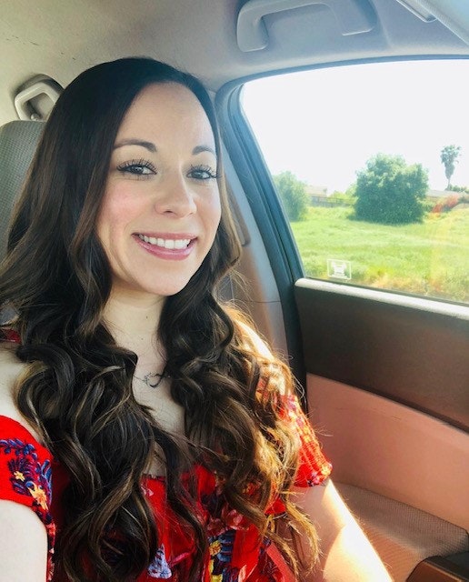 A person with long hair and a red top is smiling inside a car, with greenery visible through the window.