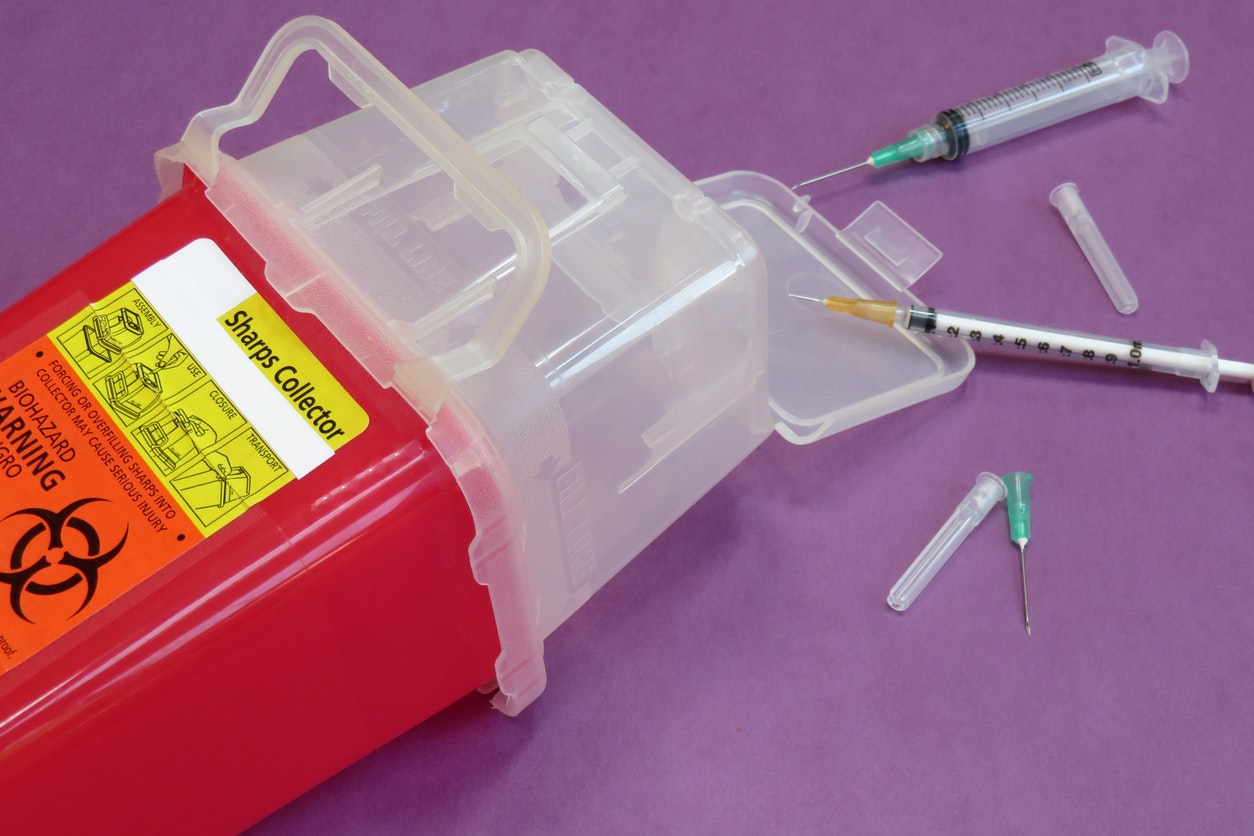 A sharps disposal container with syringes and needles on a purple surface.
