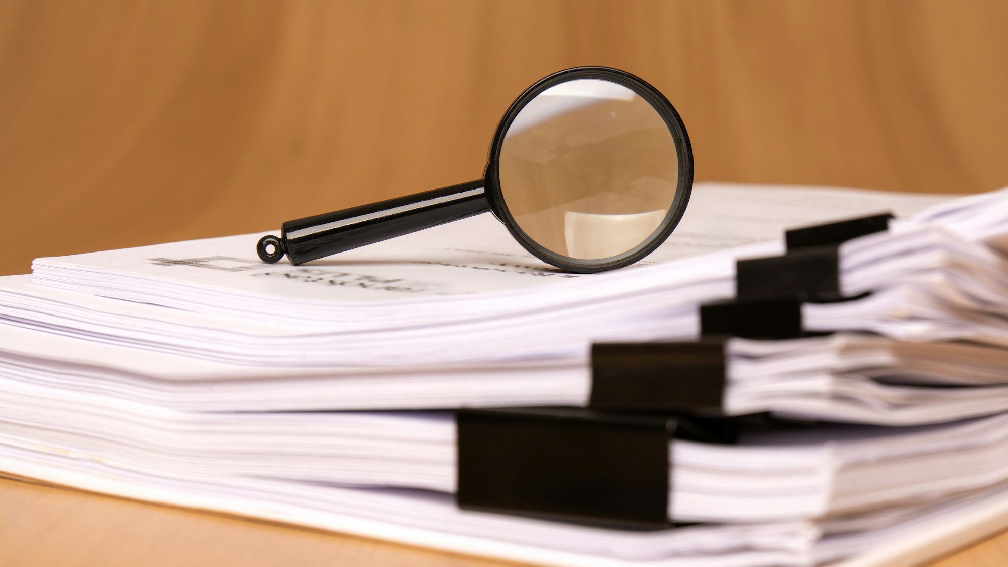 A magnifying glass rests on a stack of papers, secured with binder clips, on a wooden surface.
