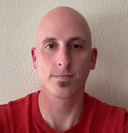 A person with a shaved head and a goatee, wearing a red shirt, against a plain wall.