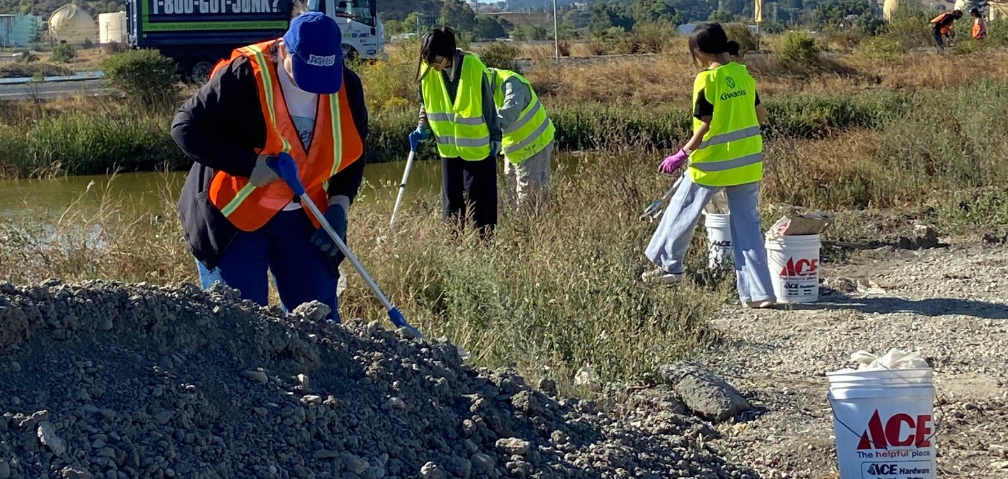 People in high-visibility vests are working or cleaning an outdoor area, with tools and buckets present.