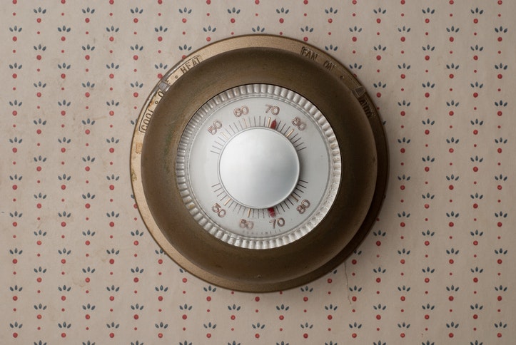 A round, vintage thermostat mounted on patterned wallpaper, set to about 70 degrees Fahrenheit.