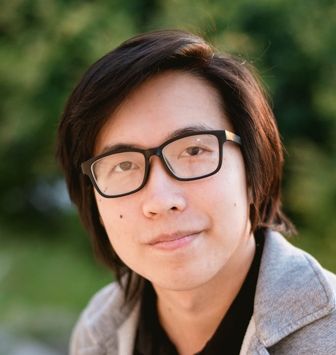 A person wearing glasses and a gray hoodie, smiling slightly with a blurred green background.