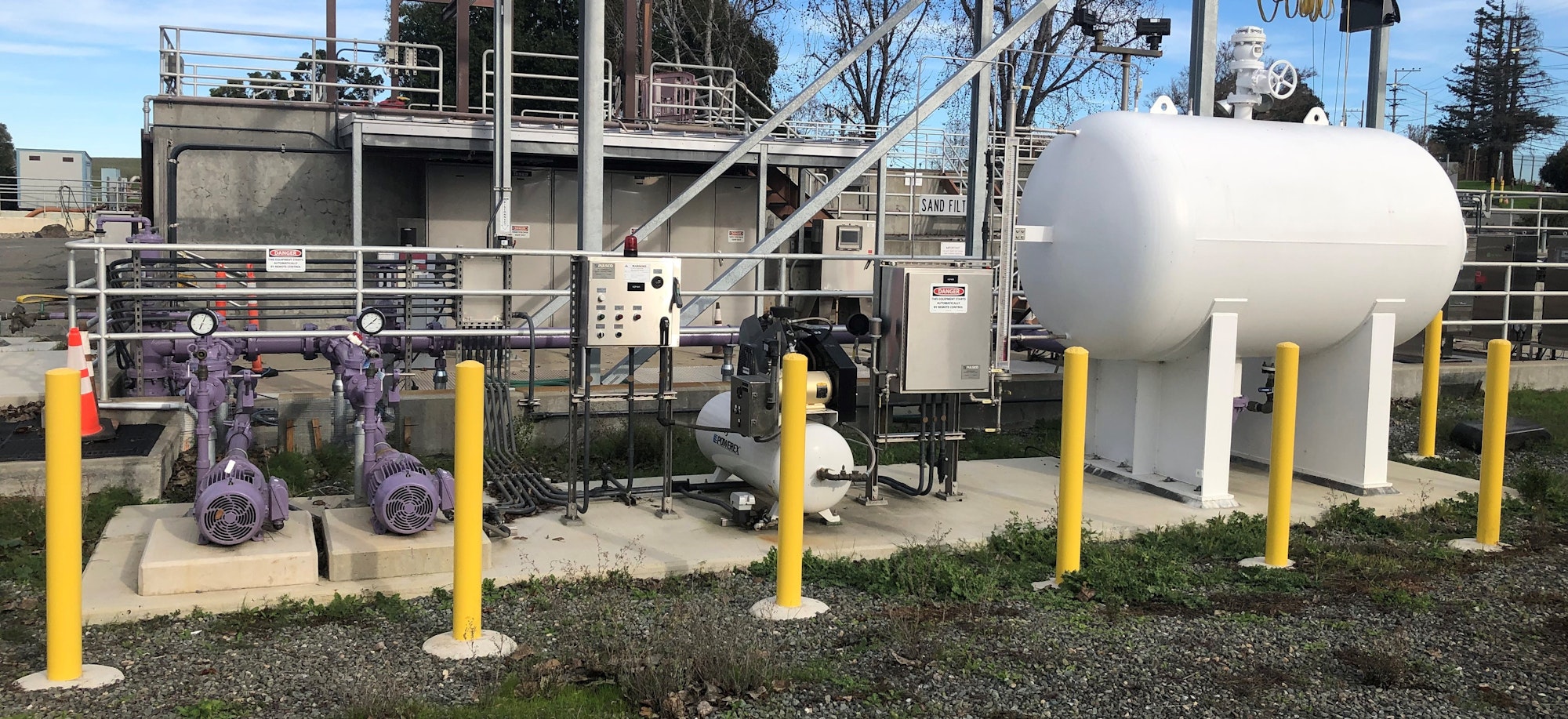 Industrial tanks, pumps, and control panels are surrounded by safety railings and posts.