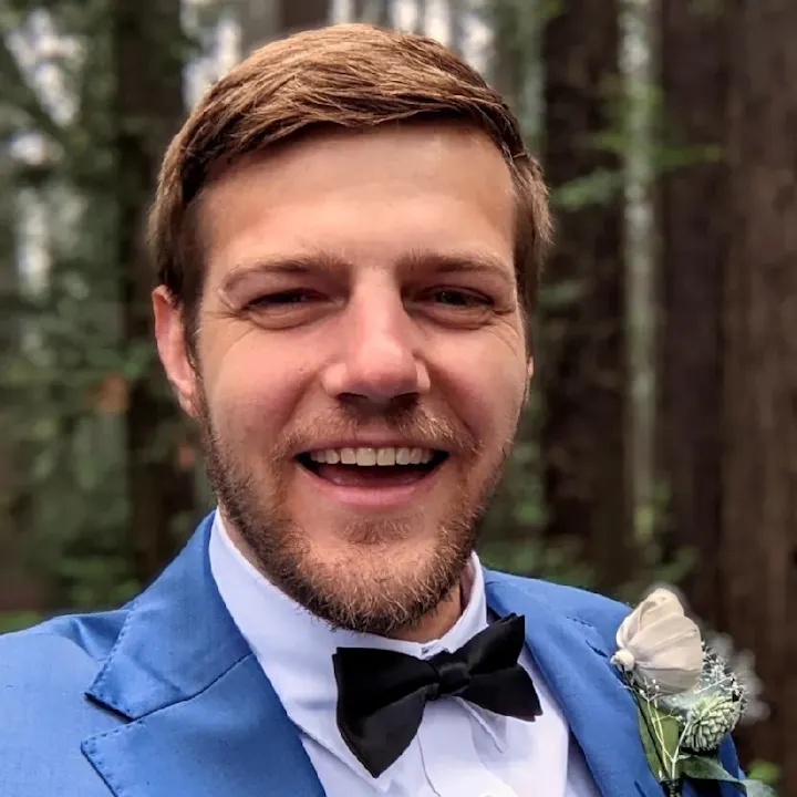 A smiling man in a blue suit and bow tie stands in a forest setting, looking happy and well-dressed for a special occasion.