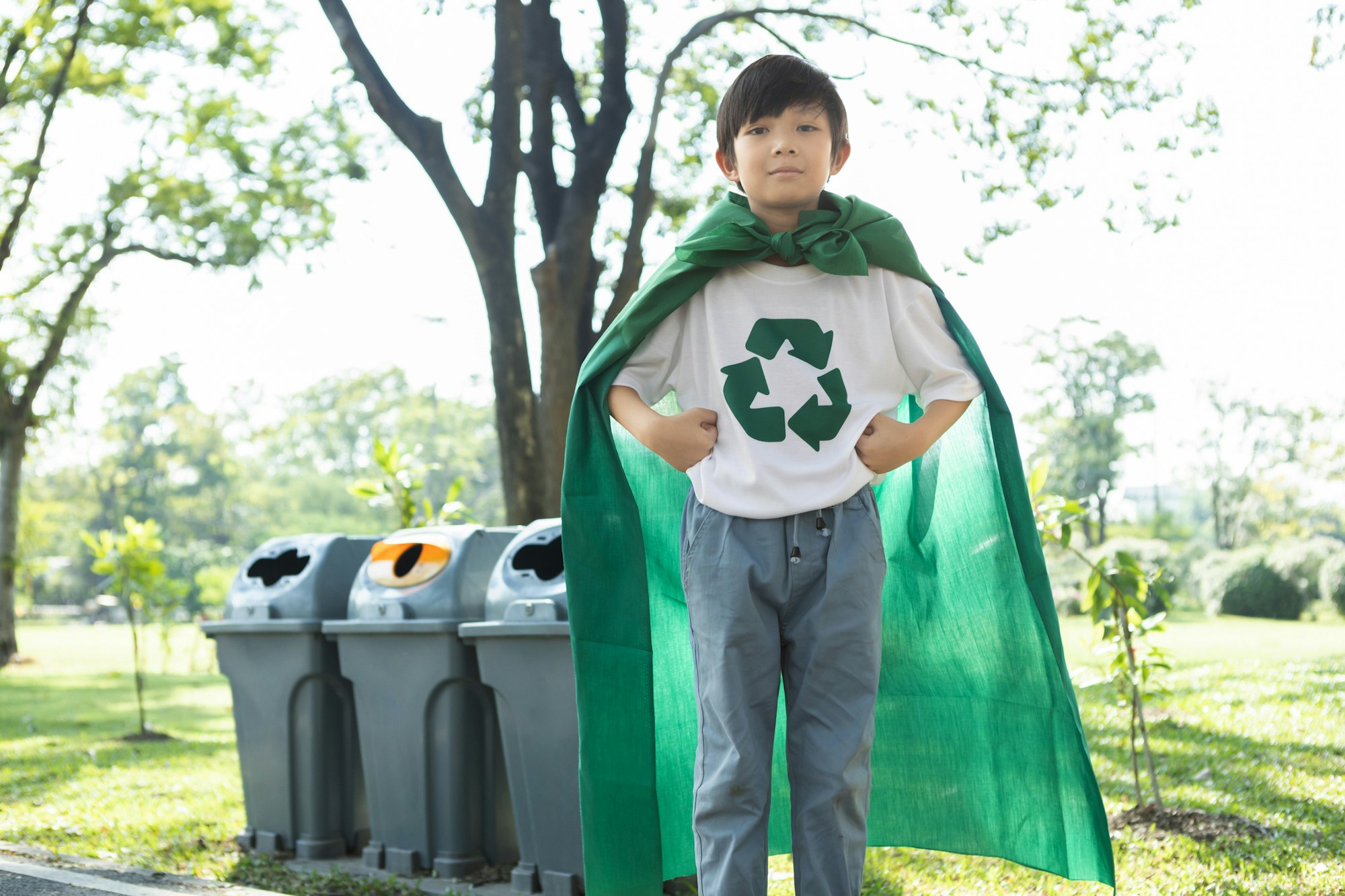 A child in a green cape and recycling shirt stands confidently near recycling bins in a park setting.