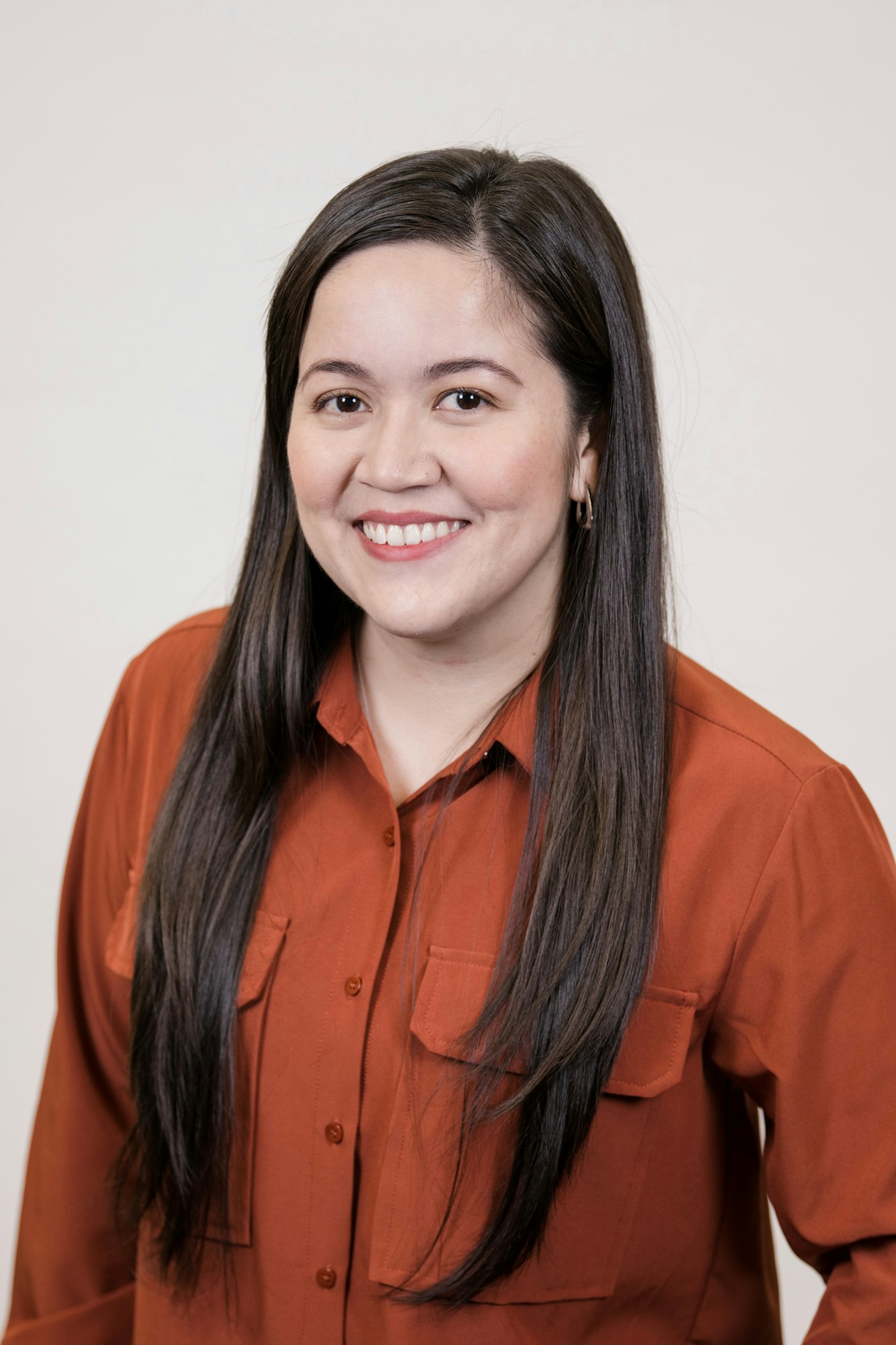 A smiling woman with long black hair, wearing an orange shirt, posed against a neutral background.