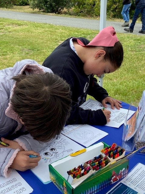 Two children are coloring at a table outdoors with coloring sheets and a box of crayons.