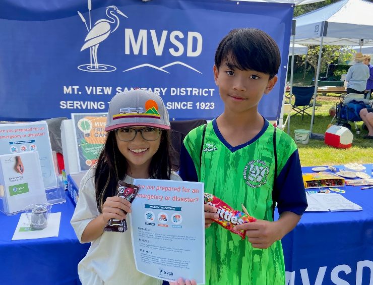 Two kids holding a flyer and snacks in front of a Mt. View Sanitary District booth at an outdoor event.