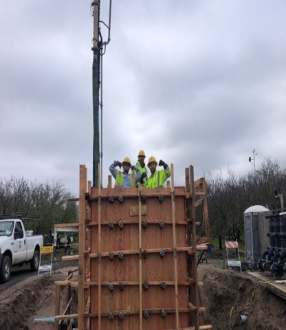 Four workers in safety gear pose by a wooden formwork structure at a construction site under a cloudy sky.