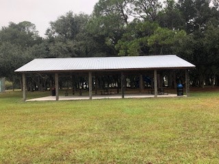 A covered picnic shelter with a metal roof in a grassy area, surrounded by trees, likely in a park setting.