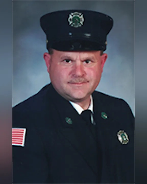 Jess Brown wearing a firefighter's uniform with a hat featuring an emblem and an American flag patch on the shoulder.