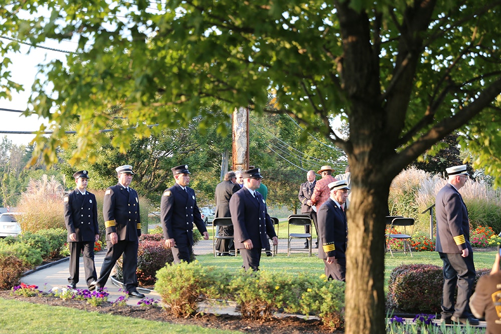 Fire, police, and other personnel at the 9-11 ceremonial event, with chairs and greenery.