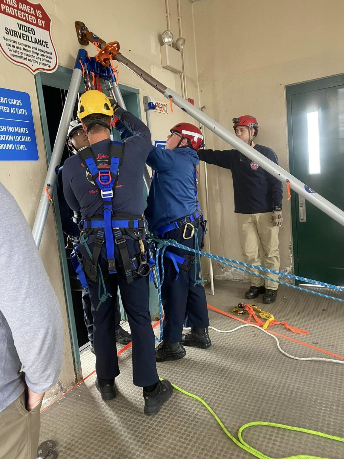 Three people in safety harnesses are engaged in a rescue training exercise indoors.