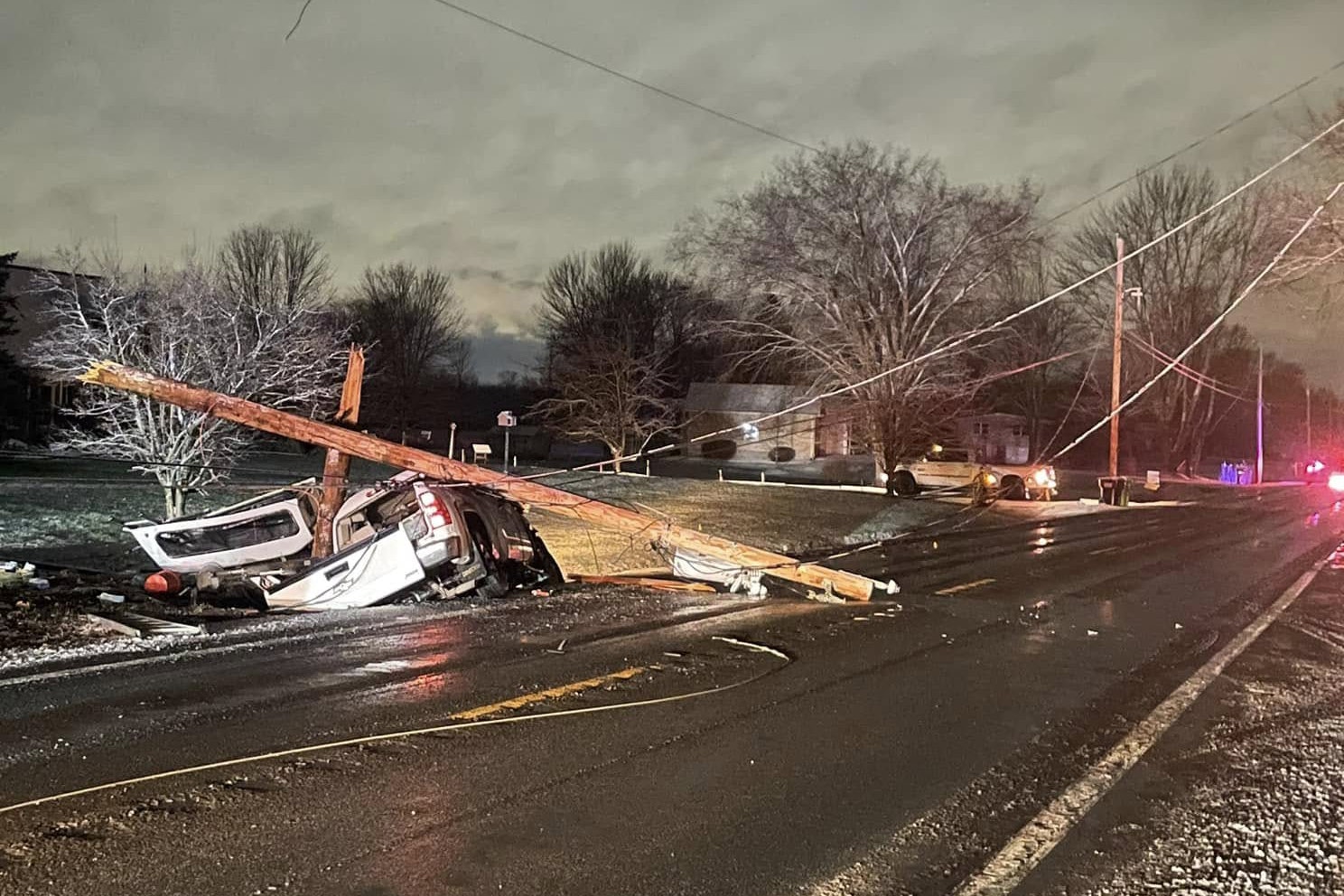 A vehicle is overturned, entangled with a fallen utility pole on a road. Emergency lights are visible in the distance.